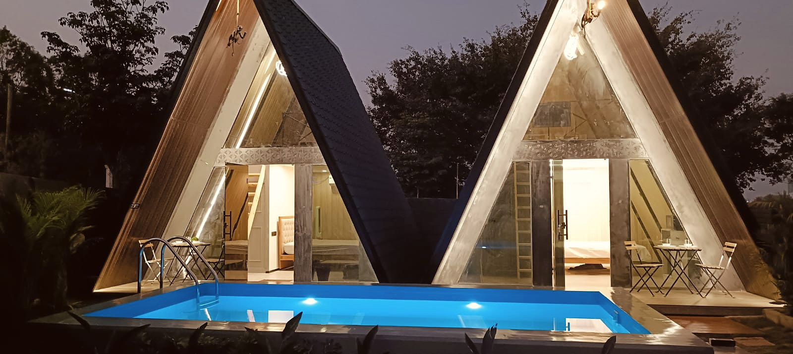 Two illuminated A-frame cottages overlooking a private blue pool at night